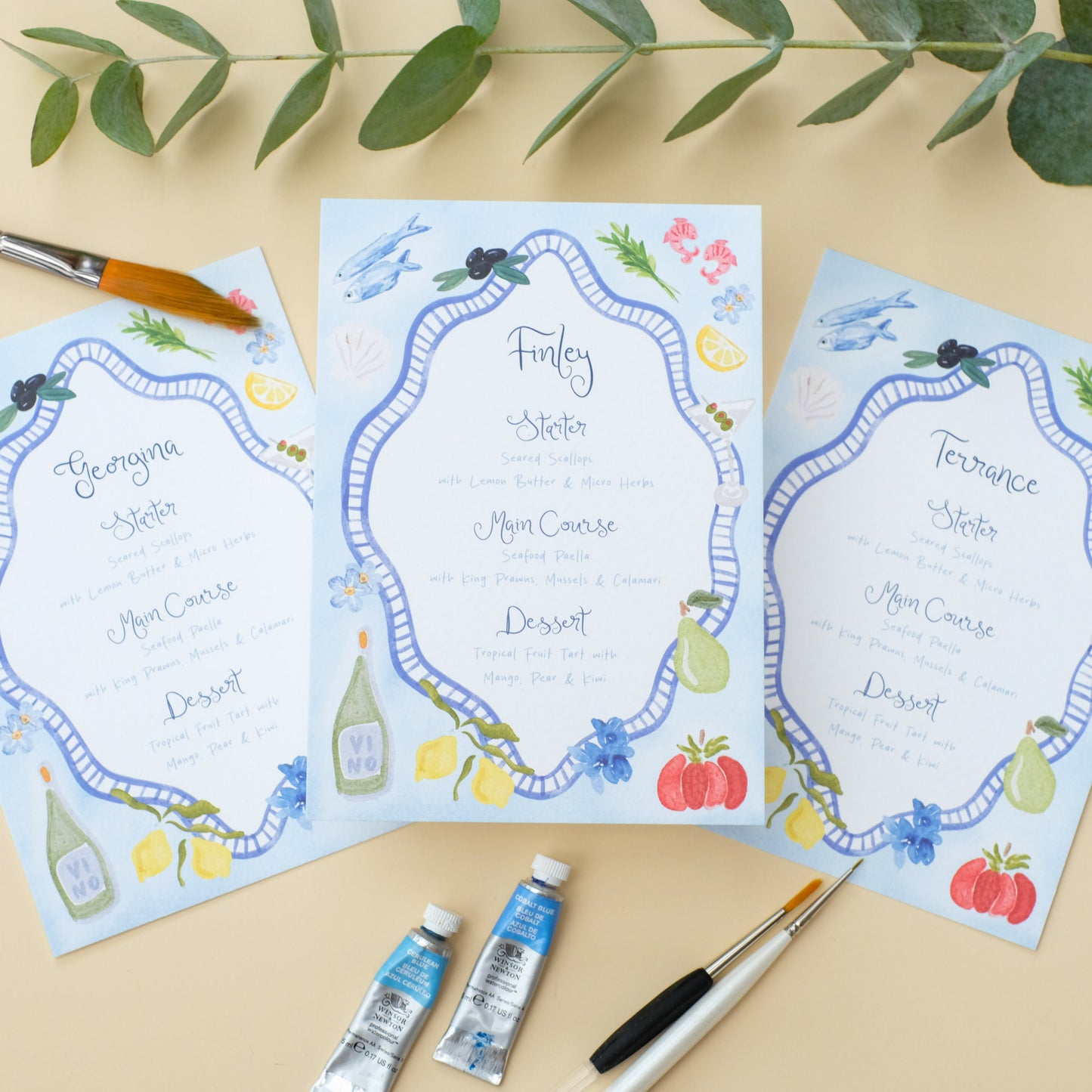 Hand painted wedding menu with blue border and watercolour illustrations of lemons, fish, shrimps and olives