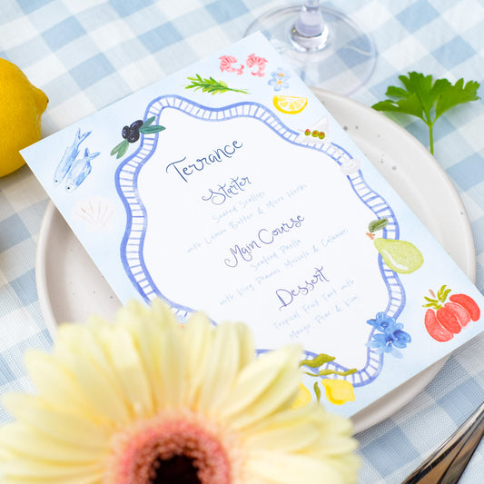Hand painted wedding menu with blue border and watercolour illustrations of lemons, fish, shrimps and olives